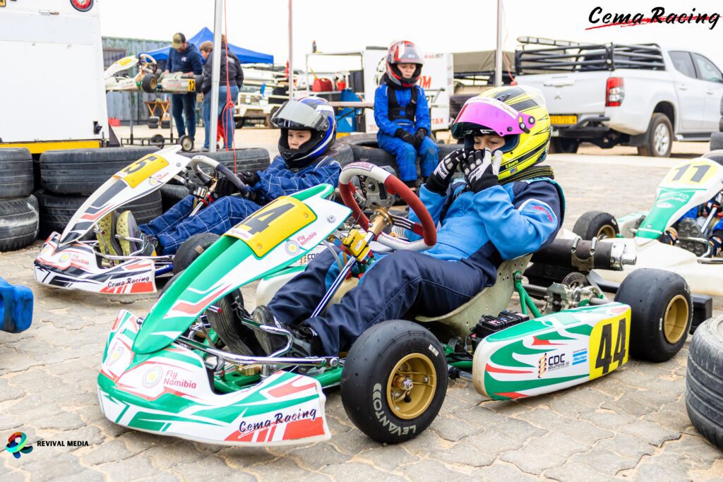 Three young kart racers preparing for a race, seated in their karts and adjusting helmets in a pit area with tires and equipment around them.