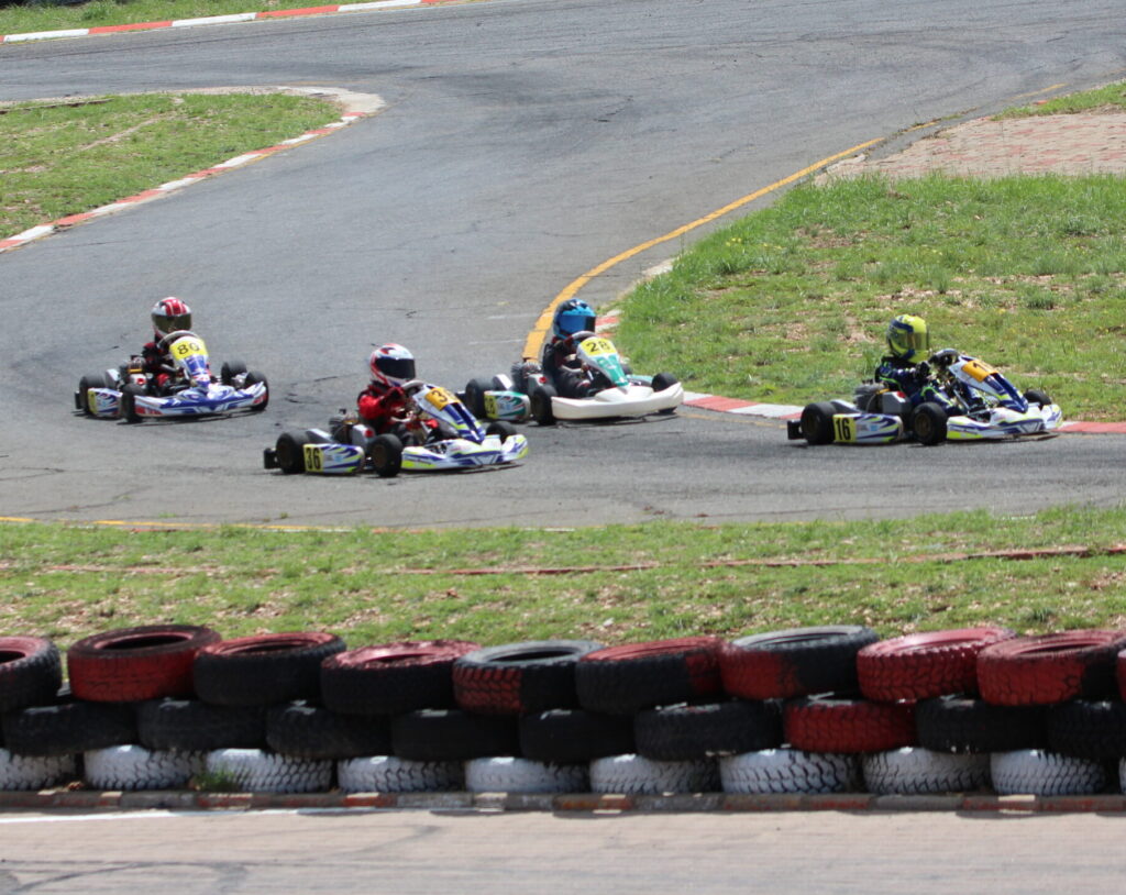 A dynamic kart racing scene on an outdoor track with four drivers navigating a curve, bordered by red and white tire barriers.