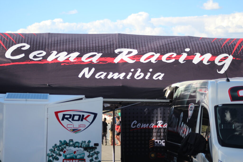 A banner reading "Cema Racing Namibia" displayed prominently above a racing team's setup area with equipment trailers at Tony Rust Race Track