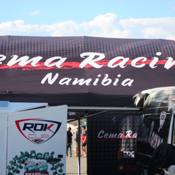 A banner reading "Cema Racing Namibia" displayed prominently above a racing team's setup area with equipment trailers at Tony Rust Race Track